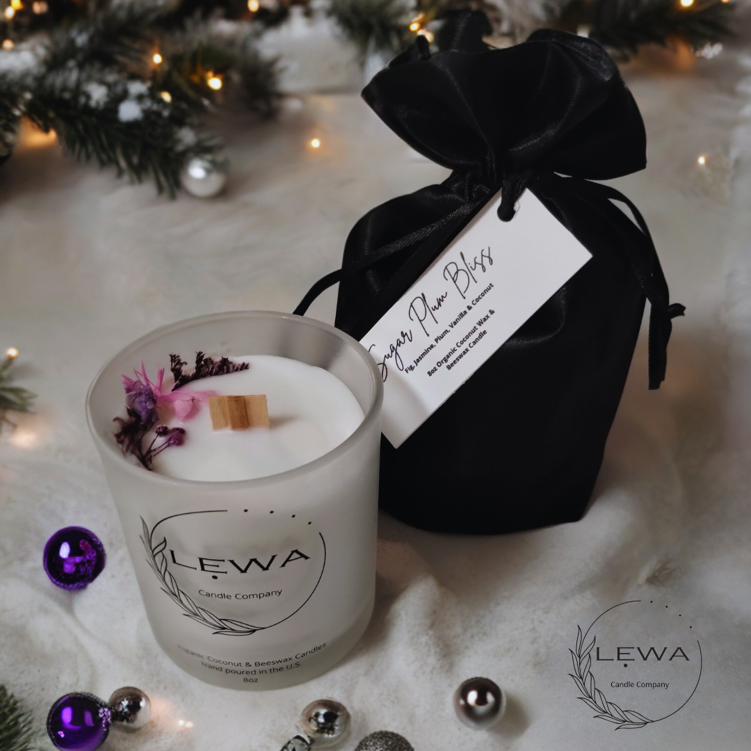 LEWA Sugar Plum Bliss candle in frosted glass jar with pink and purple floral design, presented in a luxurious black satin bag with LEWA tag, surrounded by Christmas ornaments and soft pine tree lights on a white holiday background.