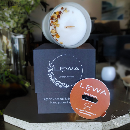 LEWA Soul candle in frosted glass jar resting sideways on signature black box with dried floral top, dust cover propped upright, displayed on a table next to a green plant and blurred living room background