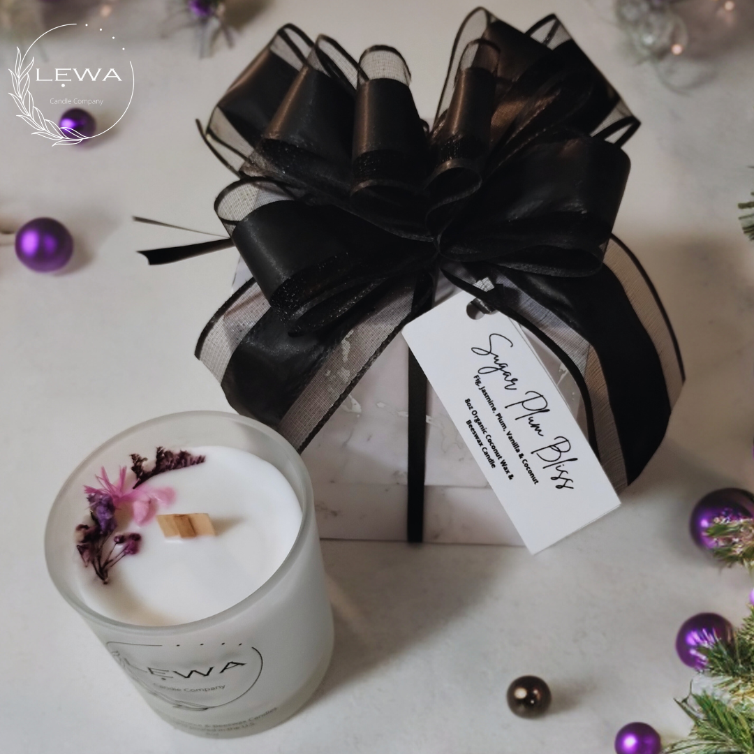 LEWA Sugar Plum Bliss candle in frosted glass jar with pink and purple floral design, packaged in a silver-white marble gift box with black bow and LEWA tag, styled on a white Christmas background with ornaments.