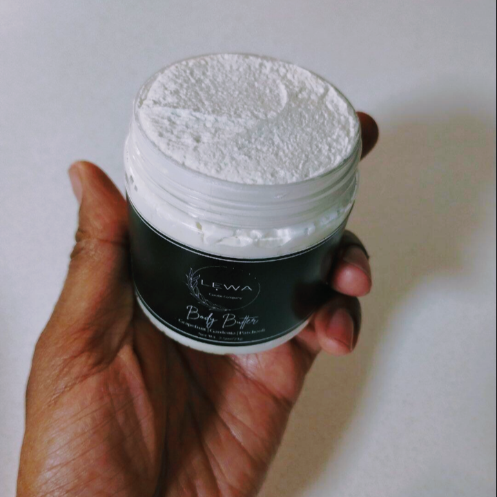 Hand holding a jar of skin nurturing 'LEWA' body butter made with organic shea, mango butter and natura oils against a neutral background.