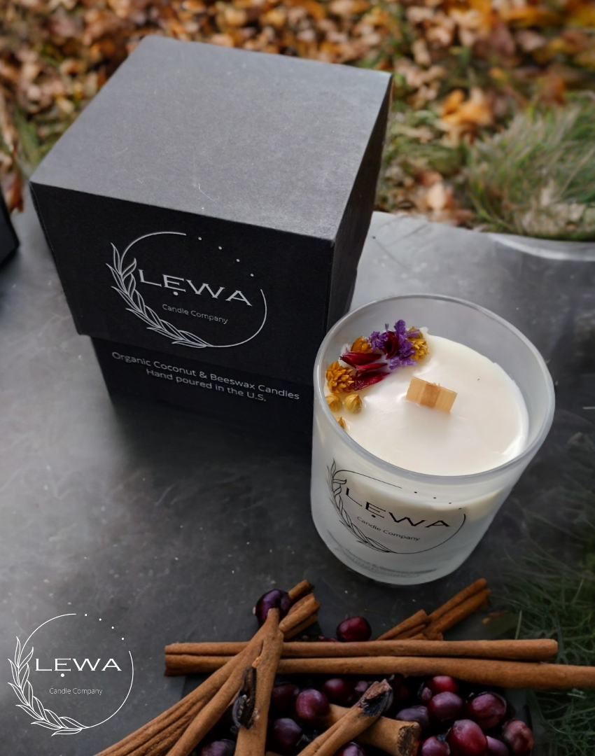 LEWA Autumn Glow candle in frosted glass jar with dried floral top, shown beside signature black LEWA box on a black outdoor table with fall leaves, cranberries, and cinnamon sticks scattered around
