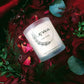 Aromatic Organic Rose Scented Candle with LEWA logo, surrounded by dried rose petals, faux roses, and greenery on red fabric.