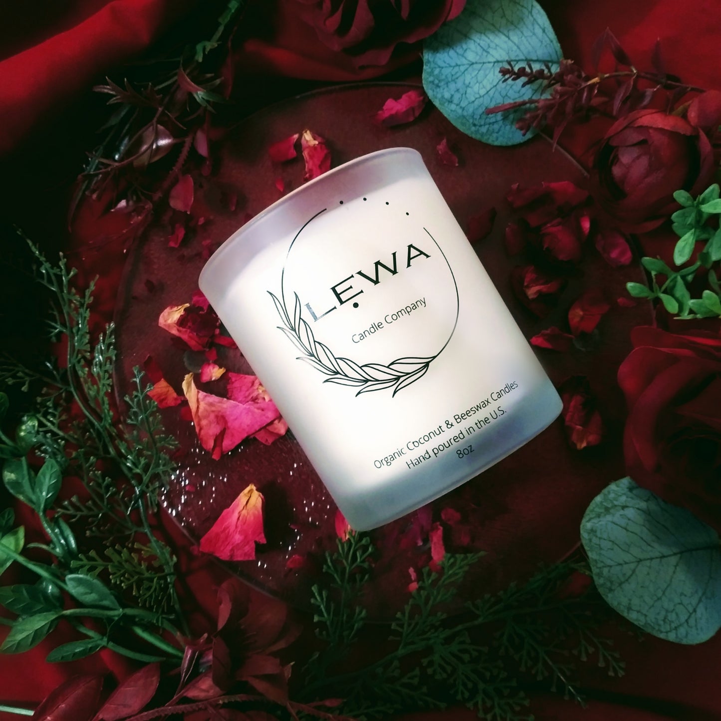 Aromatic Organic Rose Scented Candle with LEWA logo, surrounded by dried rose petals, faux roses, and greenery on red fabric.